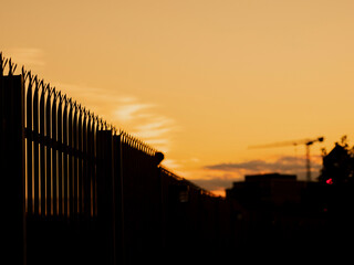 Silhouette of a metal security fence in focus. Tall construction crane in the background. Warm orange sunset sky. Abstract city background.
