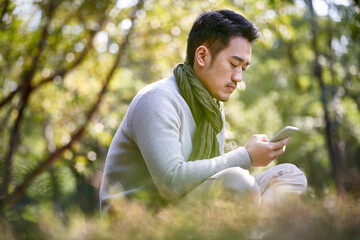 young asian man sitting on grass in park looking at cellphone