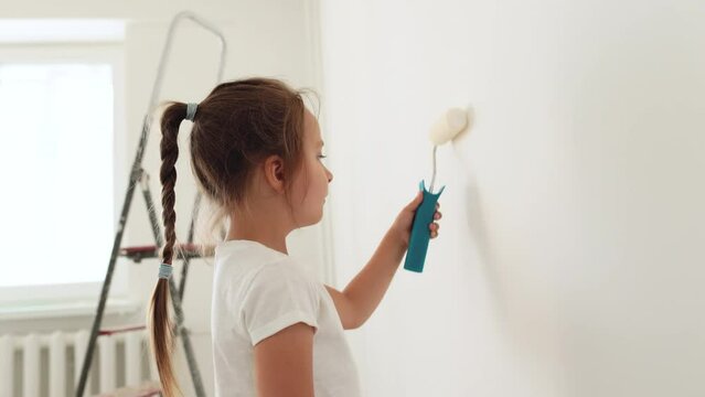 Repair in the apartment. Happy cute child girl painting the wall with paint roller standing in a bright room. Home repairs concept.