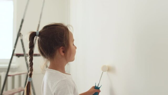 Cute little toddler girl painting the wall with white color in new house. Family repair apartment home. Happy child girl paints the wall, having fun with brush, indoors