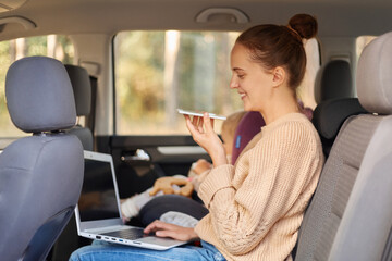 Side view portrait of woman working on laptop and using voice assistant for searchng information while sitting with her baby daughter in safety chair on backseat of the car.