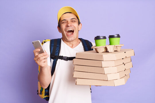 Extremely happy man courier in yellow cap and T-shirt holding food order pizza boxes and coffee to go, standing with phone in hands, screaming happily, isolated on purple background.