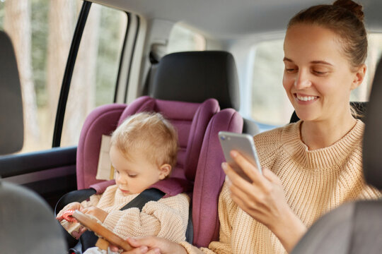 Image of happy satisfied delighted mother using phone while sitting in car with her toddler daughter on backseat, female looking at smart phone screen, checking social networks.