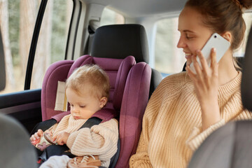 Portrait of smiling attractive mother using phone while sitting in car with her infant daughter on...