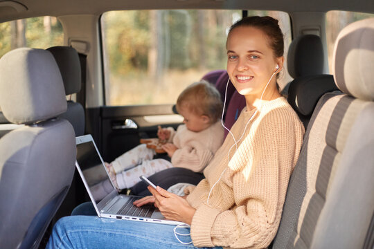 Portrait of smiling woman sitting with her baby daughter in safety chair on backseat of the car, working on laptop, going to office and kindergarten, looking at camera with happy expression.