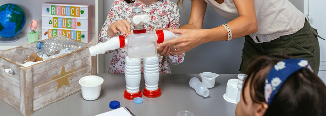 Teacher in ecology classroom helping to girl make recycled toy robot with plastic packages