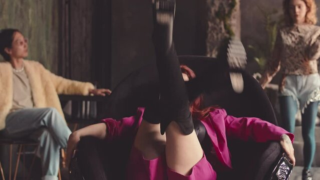 Slowmo of young diverse people in glamourous 80s style outfits vogue dancing in ethnic studio. Red haired woman in pink performing eccentric leg movements for camera sitting on chair