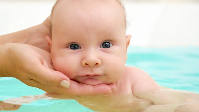 Mother teaches a newborn baby to swim and stay on the water. Two-month-old baby is bathing in a large bathtub. Newborn baby is in the water, bathroom. Hygiene procedures of a newborn baby