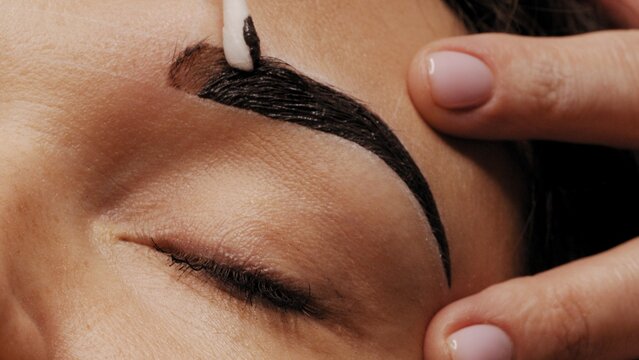 Cosmetologist removes black eyebrow paint after coloring. Professional eyebrow coloring in black in a beauty salon. Working hands of the master close-up. Beauty treatment procedures