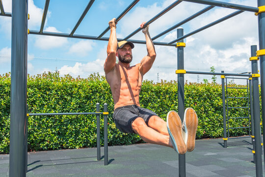 Male athlete in a cap without a T-shirt, muscle training, abdominal exercises, leg raises on the bar. In the summer in the city on an open sports ground. Active lifestyle, workout, outdoor fitness.