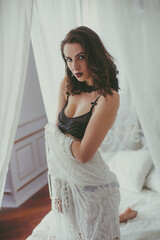 Fototapeta premium A young attractive girl with dark hair in black lingerie with evening make-up and jewelry and a white bathrobe on a white bed in the bedroom. beauty and fashion