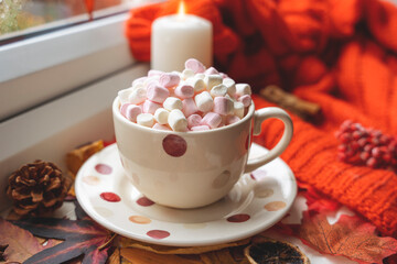 Obraz na płótnie Canvas a polka dot marshmallow cup with a saucer on autumn leaves with an orange scarf a candle a cone on the windowsill in rainy weather