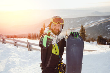 Happy Man snowboarder stands on top of mountain background ski resort sunset. Concept banner winter...