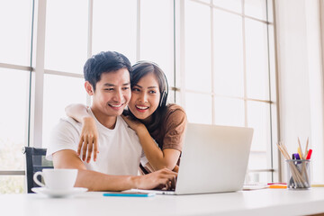 Young Asian couple playing laptop and looking at the screen at home. Cheerful couple relaxing in beautiful white room. Close up shot woman hugging her boyfriend while he working.