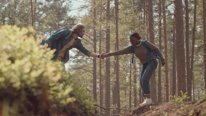 Tracking shot of young diverse couple with backpacks hiking in forest at summertime. Boy helping girl to jump through rough area - Powered by Adobe
