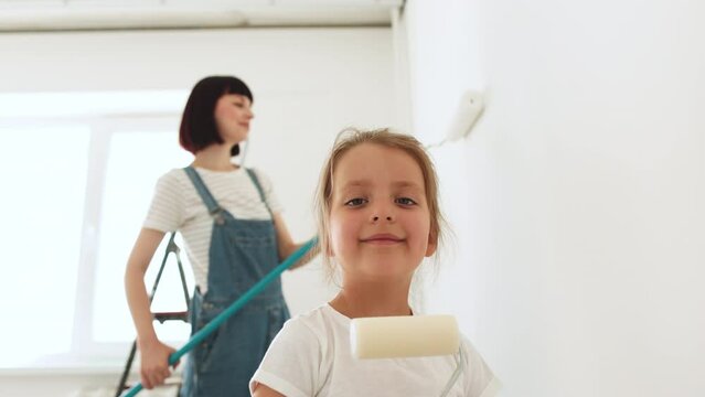 Young family of beautiful mother and cute little daughter having fun together while repairing room. Little girl with paint roller coloring with mom the wall. Focus on daughter.