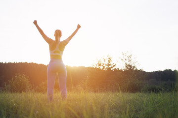 a girl in a fitness suit stands with her back to the camera at sunset with her hands raised after an outdoor workout, a girl silhouette motivational place for an inscription. High quality photo