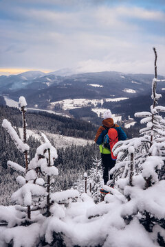 Candid portrait of young smiling man wearing colourful winter hiking jacket, warm-ups and white cap overlooking the snowy area of Stare hamry with the highest point of the Beskydy Mountains Lysa hora
