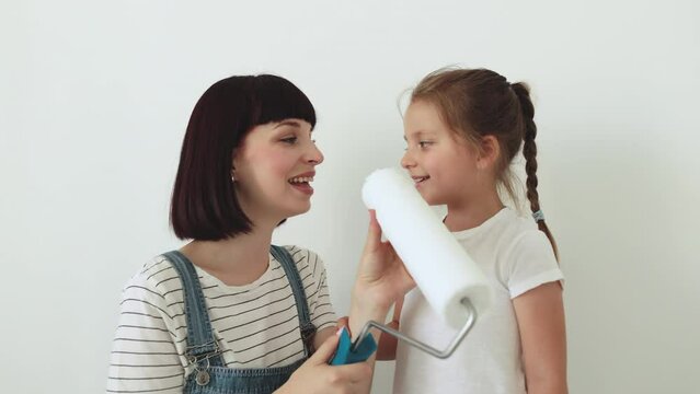 Happy family, young caucasian mother and her cute child daughter singing and paints the wall with white paint. Repair in the apartment concept.