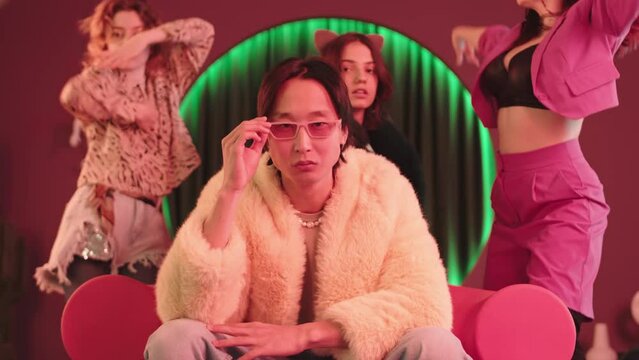Slowmo of Asian hipster boy in trendy pink eyeglasses and fur coat looking pompously at camera while three attractive girls in fancy outfits vogue dancing behind in retro studio with neon pink light