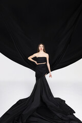 Fashion, fine art, fantasy concept. Beautiful brunette woman with red lips wearing huge black dress with flying trails like wings in white background with copy space. Image with motion blur