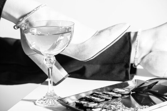 Fashion and style concept. Fancy accessories, casino tokens, sunglasses, jewelry and drink placed on table. Woman legs with black trousers and high heel shoes in background. Black and white image