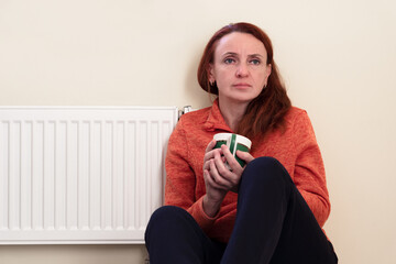 A woman with a cup of hot tea is heated by a radiator. Energy crisis. Depressive mood. Age crisis.