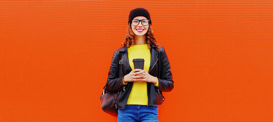 Portrait of stylish smiling young woman with cup of coffee wearing black rock style leather jacket, hat, eyeglasses on red background