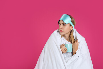 Sad young woman in sleeping mask wrapped with blanket holding cup on pink background. Space for text