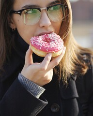 A young girl with glasses takes a bite of a glazed donut. Selective focus on the donut. The girl is wearing a coat