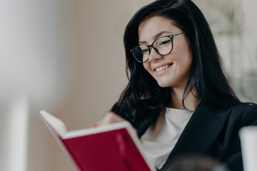 Cheerful dark haired woman creats do list with targets, holds notebook, writes organisation plan, wears spectacles, dressed in formal outfit, smiles pleasantly. Autodidact and education concept