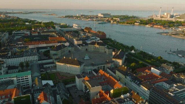 Fly above historic building at waterfront at golden hour. Aerial view of Amalienborg palace with equestrian statue on square. Copenhagen, Denmark