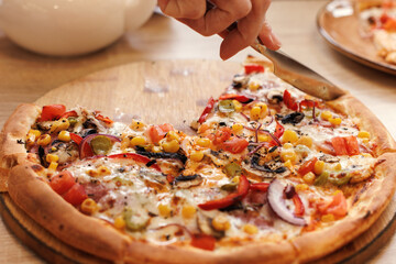 A piece of pizza is cut off with a table knife on a round cutting board. Selective focus.