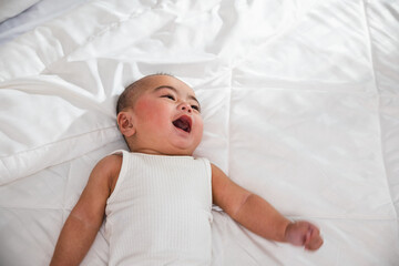 Asian cute little baby on bed under soft blanket, baby wearing bodysuit lying on white beedsheets...