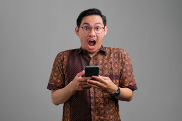 Shocked young Asian man wearing batik shirt using smartphone and looking at camera isolated on grey background