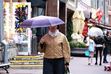 Rain in autumn city, woman with umbrella wearing jacket walking on а street on people and shops background