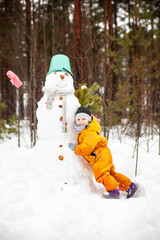 Three-year-old girl in  orange jumpsuit with  snowman outside in winter