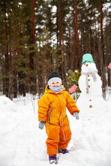 Fototapeta na wymiar Three-year-old girl in orange jumpsuit with snowman outside in winter