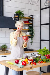 beautiful elderly gray haired senior woman cook in cozy kitchen with fresh organic vegetables, tomatoes, cabbage, lettuce, cucumbers on table cooking healthy vegetable salad, healthy food active life