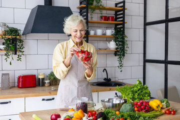 beautiful elderly gray haired senior woman cook in cozy kitchen with fresh organic vegetables, tomatoes, cabbage, lettuce, cucumbers on table cooking healthy vegetable salad, healthy food active life
