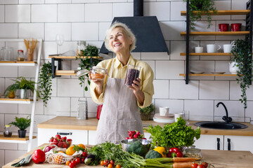 beautiful elderly gray haired senior woman cook in cozy kitchen with fresh organic vegetables, tomatoes, cabbage, lettuce, cucumbers on table cooking healthy vegetable salad, healthy food active life