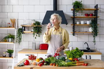 beautiful elderly gray haired senior woman cook in cozy kitchen with fresh organic vegetables on...