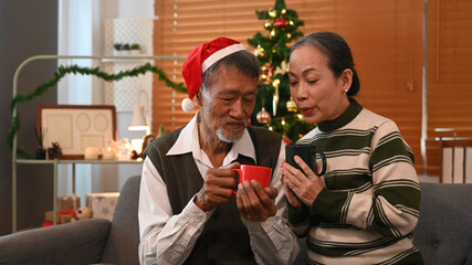 Happy senior couple sitting on couch next to a decorated Christmas tree and enjoying drinking hot chocolate together