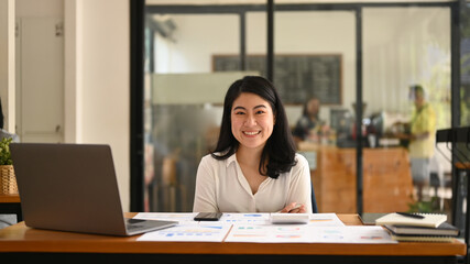Obraz na płótnie Canvas Cheerful asian female employee sitting with arms crossed at her office desk and smiling at camera