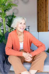 happy beautiful elderly gray haired senior woman sit on sofa in living room in cosy apartment, smiling elderly woman, modern granny candid portrait