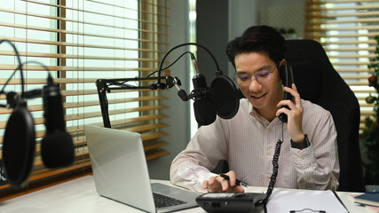 Asian man radio host talking on telephone and using condenser microphone recording voice conversation at home studio