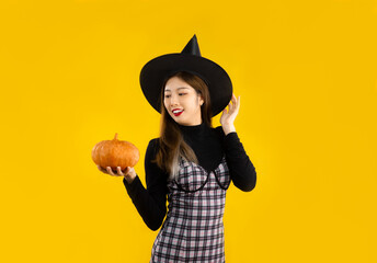 Halloween theme, young asian girl in witch costume holding orange pumpkin posing on yellow color background.
