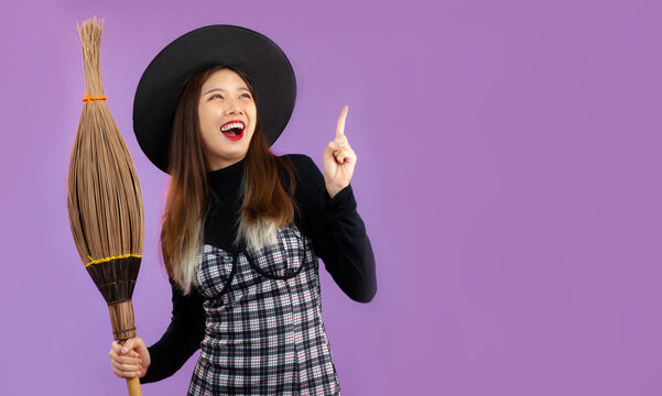 Halloweeen theme, young pretty asian woman in witch costume holding broom posing finger pointing on purple background.
