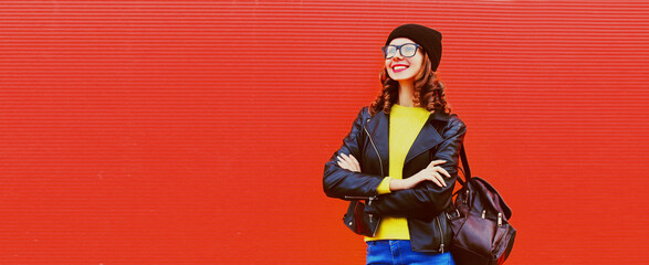 Portrait of stylish smiling young woman posing wearing backpack, black rock style leather jacket, hat, eyeglasses on red background