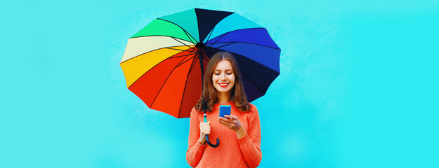 Autumn portrait of happy smiling young woman calling on smartphone with colorful umbrella on blue background